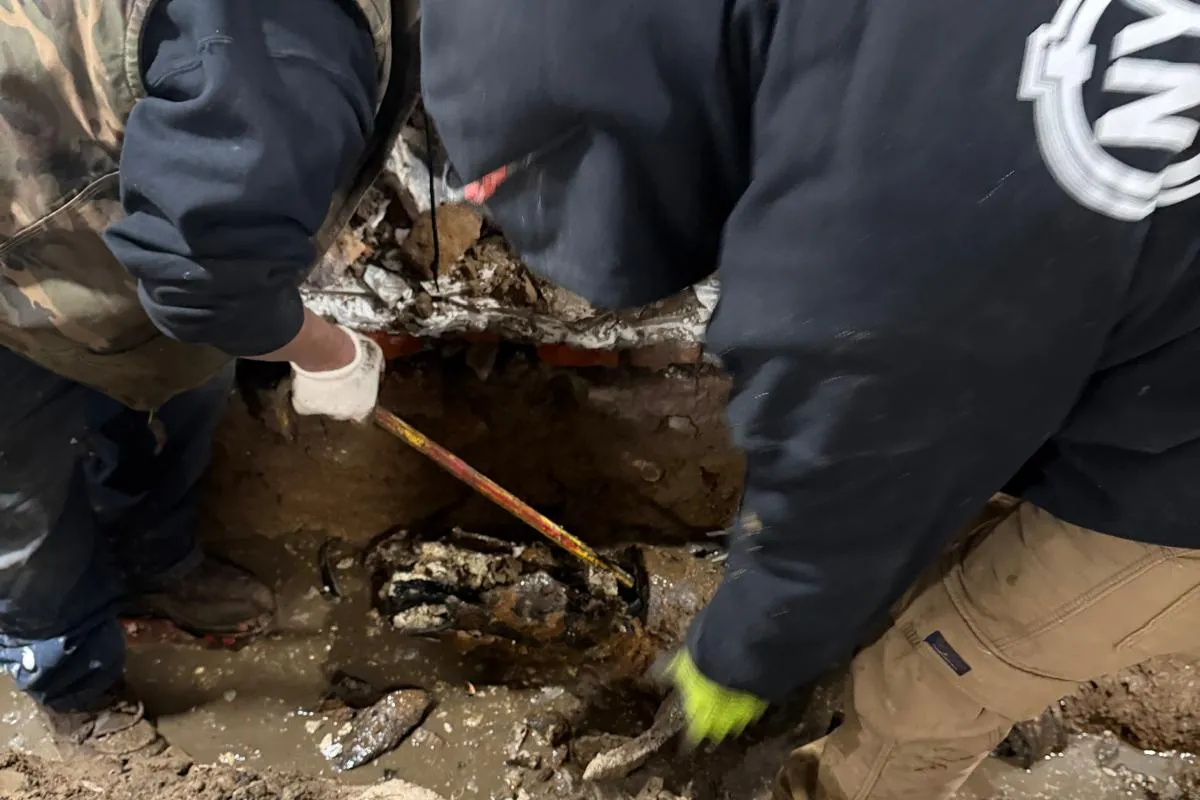 Two NY Sewer and Drain technicians in branded gear excavating a broken cast iron line in a New York City basement